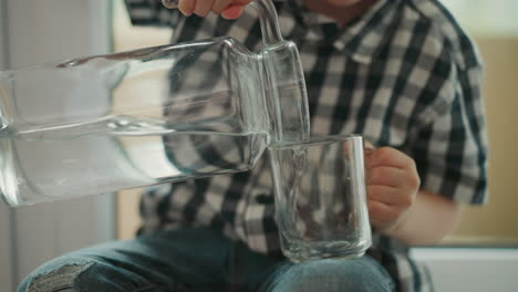 close up of young boy wearing checkered shirt carefully pouring water from large clear jug into glass cup placed on his leg while sitting indoors with soft daylight illuminating scene