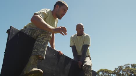 two diverse fit male soldiers helping another climb high hurdle on obstacle course