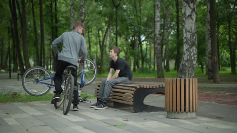 young boy sits on park bench with his head bowed, his bicycle placed upside down beside him, another boy approaches riding a bicycle, and they begin a conversation in a serene park