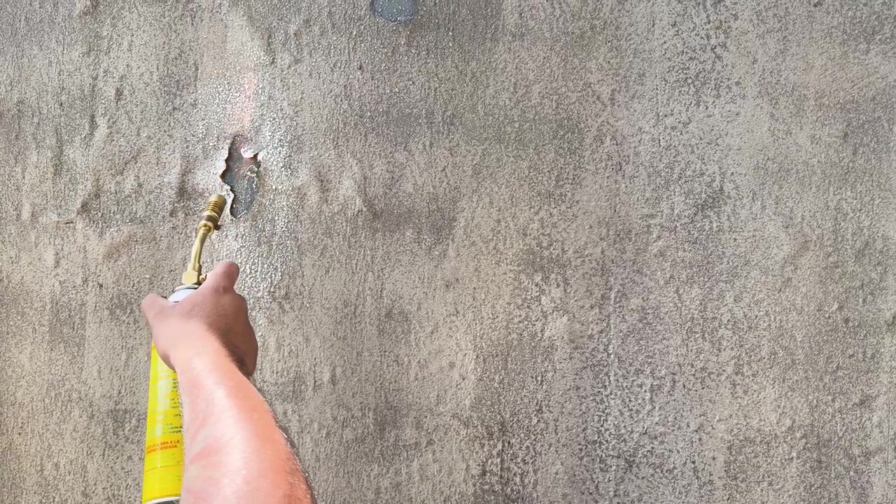 Hand uses blowtorch to create rough textures on gray plaster wall during surface work, lifts and spreads flame to cause paint to bubble