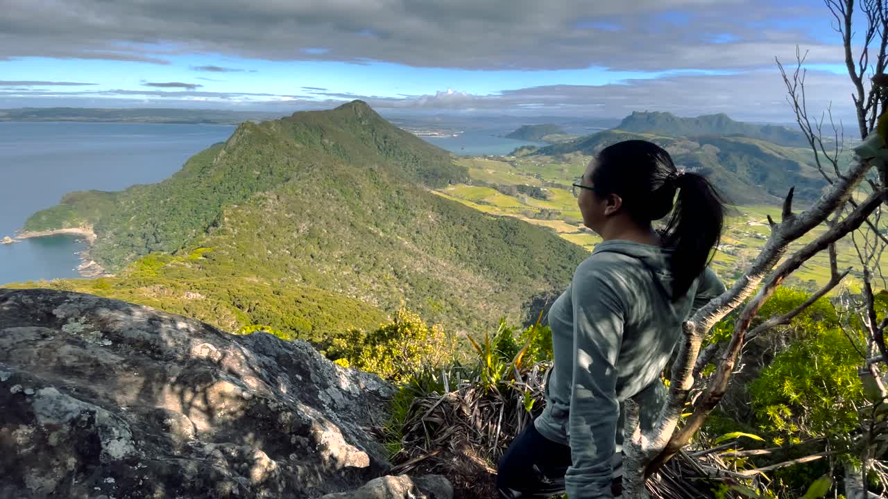 회색 스웨트 셔츠를 입은 어두운 머리 여성은 바람이 불고 있는 절벽 꼭대기에 서서 울창한 산과 바다를 바라보고 있습니다.