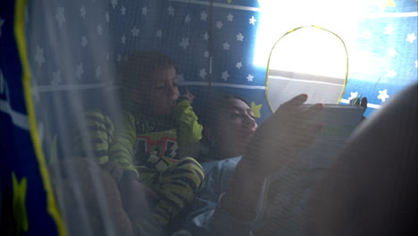 Slow-motion-of-a-young-mexican-latin-mother-reading-a-book-to-her-son-inside-his-blue-castle-tent-wearing-pajamas-enjoy-a-time-together