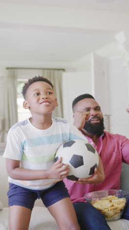 vertical video of african american father and son watching match and supporting, in slow motion