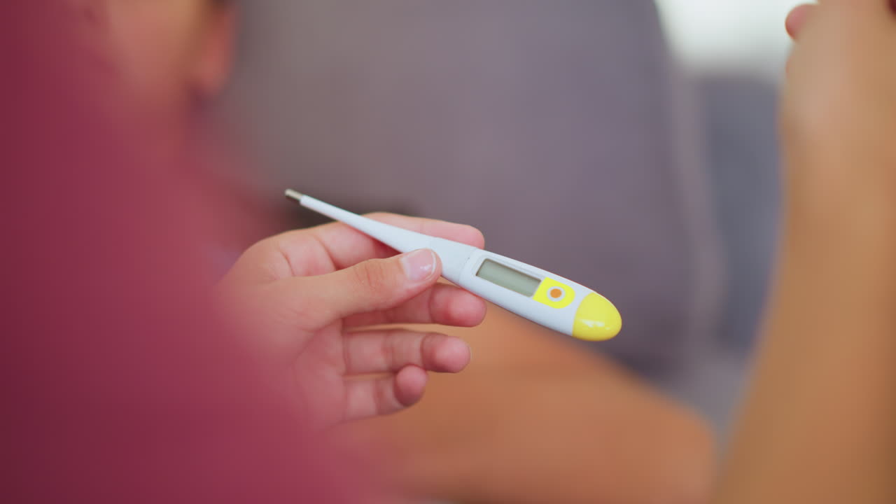 Close up of girl in maroon shirt raising hand while observing thermometer with calm serious expression as sick person lies blurred in background