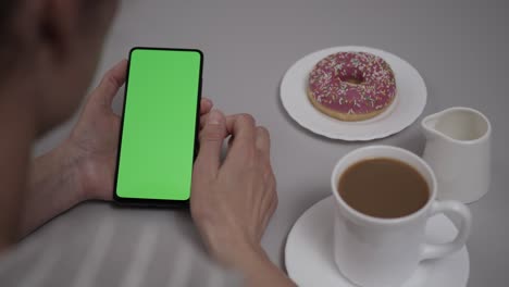 woman sitting at table coffee donut using smartphone with chroma key green screen, scrolling through social network media online shop internet. smartphone in horizontal mode with green screen mock-up.