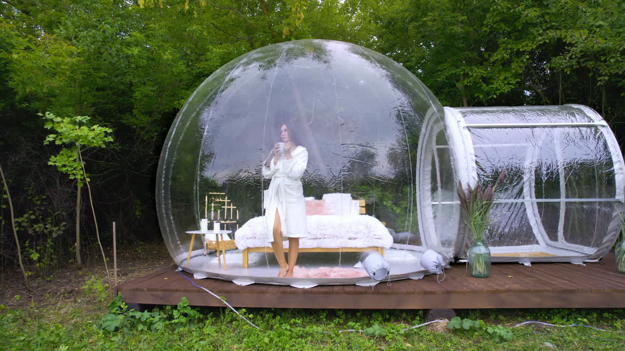 Gennemsigtigt bobletelt ved glamping, frodig skov rundt om og indvendigt. Kvinde med en kop, der kigger ud.