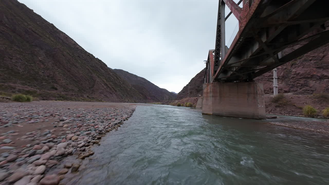 FPV drone sweep captures the dramatic steel truss bridge spanning the Las Cuevas River in the Andes near Mendoza, Argentina, built as part of the legendary Ferrocarril Trasandino -Transandine Railway