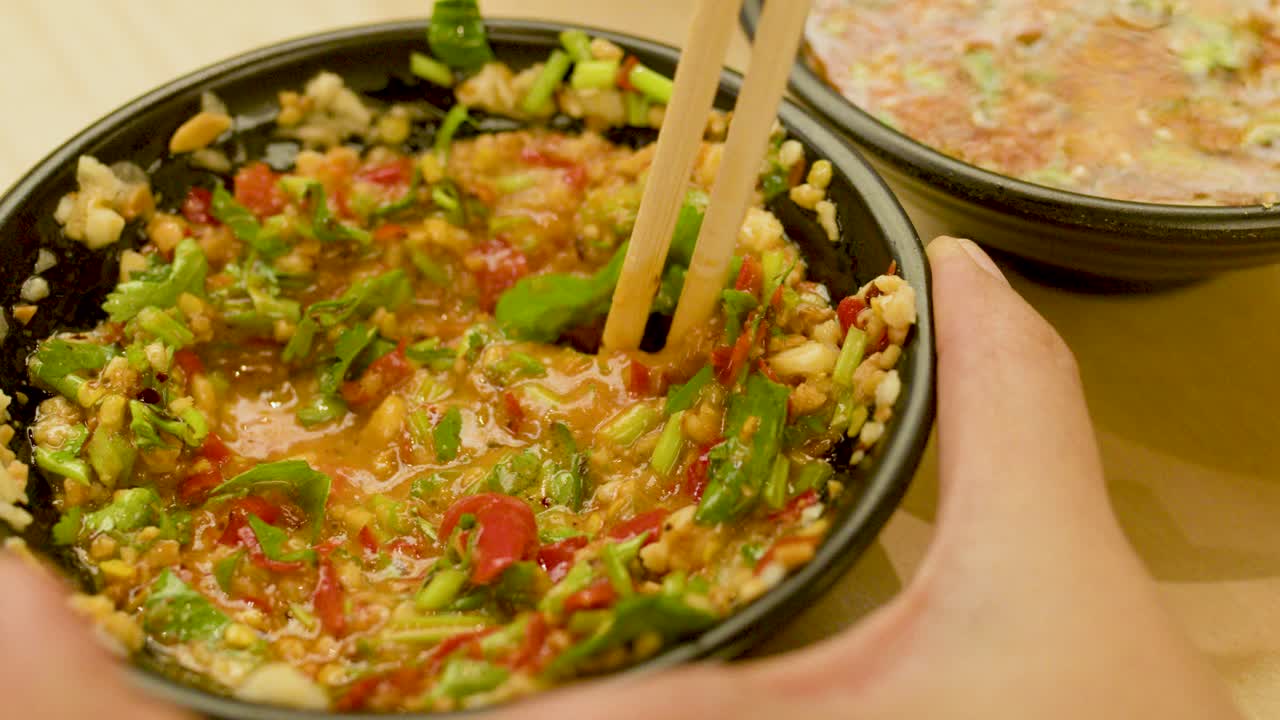 Hands use chopsticks to mix spicy rice, chili, coriander, and garlic in warm indoor lighting