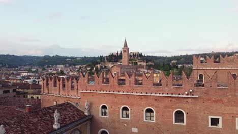 Drone aerial of Verona shows the brick Gothic Palazzo del Podestà with its battlements, and the tall bell tower of San Zeno in the distance, surrounded by hillside cypresses, slow glide, wide shot