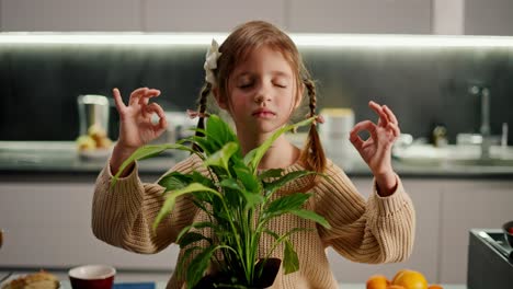 Close-up-of-a-small-calm-brown-haired-girl-with-a-braided-hairstyle-sits-in-the-lotus-position-and-practices-Zen-and-meditation-and-holds-a-houseplant-in-front-of-her-in-a-modern-kitchen-in-the-evening