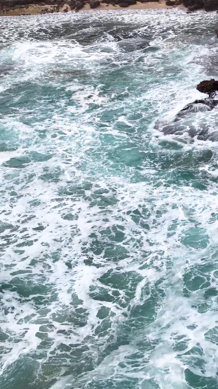 Onde turchesi turbolente si infrangono contro rocce e una riva sabbiosa, catturate in piena luce naturale, vista aerea.
