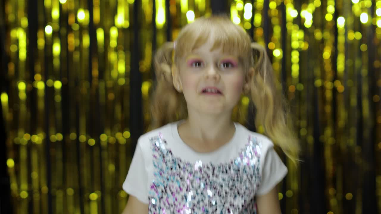 행복한 아이는 엄지손가락을 올리고 미소 짓고 카메라를 쳐다보고 있습니다. 소녀는 포일 커튼과 함께 배경에 포즈를 취하고 있습니다.