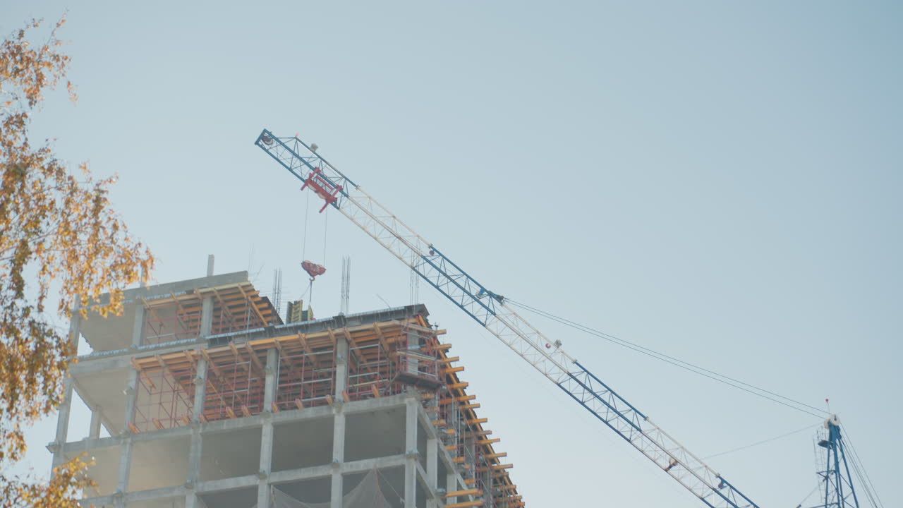 Crane cluster over rising concrete frame, flags and autumn foliage in foreground, close view of top floors with hoist cables and scaffold, clear blue sky, dense urban construction energy