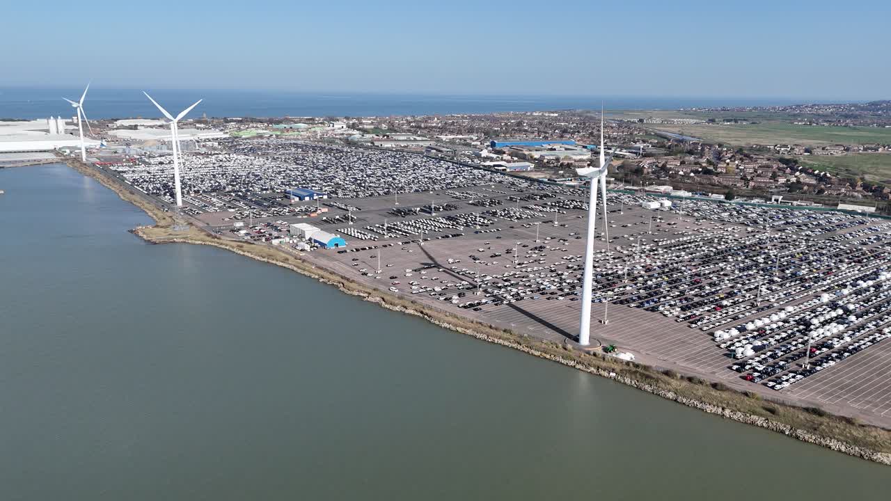 Hilera de turbinas eólicas con coches de importación nuevos aparcados en Sheppey, Kent. Grabación con dron, aérea.