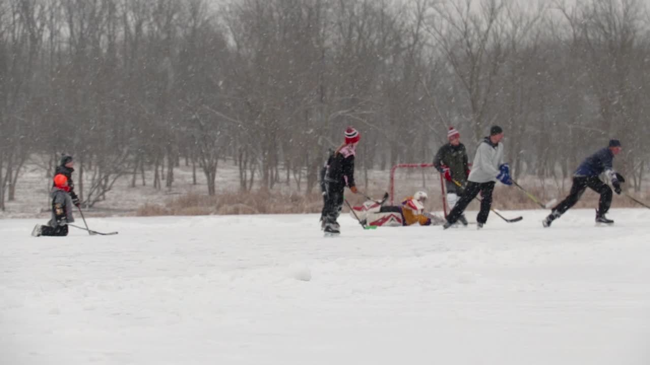 Wide angle following shot of group of teenagers, children and adults playing pond hockey. They are playing on a frozen lake and it's snowing a lot.