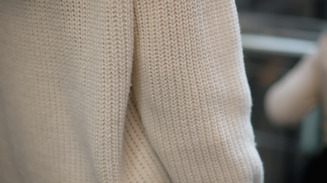 back view of refined shopper from head to waist on moving walkway in mall, pastel hair in white knit sweater holding shopping bag, motion blur capturing ambient lights and travelator rails