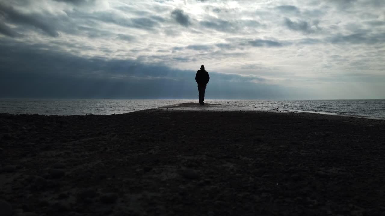 Man in a black coat walking on a concrete jetty to the infinite horizon of Lake Ontario