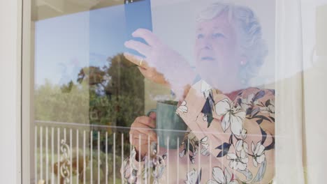 Kaukasische-ältere-Frau-Hält-Eine-Kaffeetasse-In-Der-Hand-Und-Macht-Ein-Foto-Vom-Smartphone-Durch-Das-Fenster