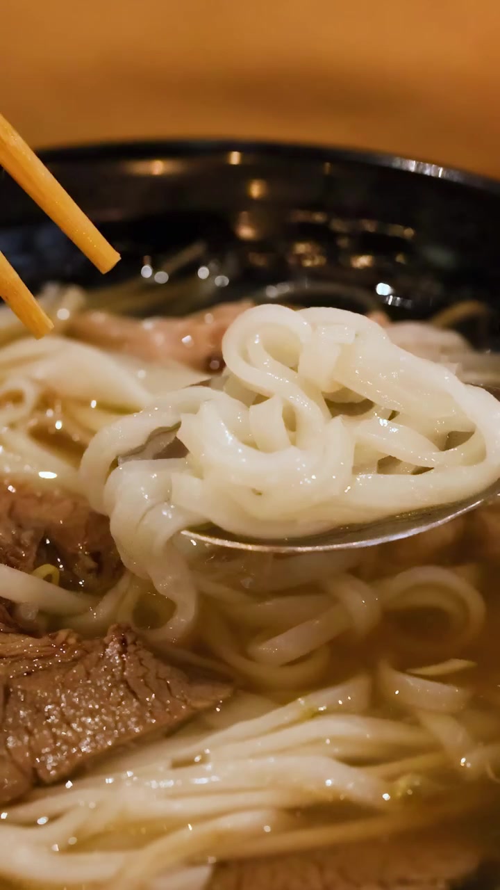 Un primo piano del pho vietnamita gustato con bacchette e un cucchiaio, che evidenzia i noodles e il brodo.
