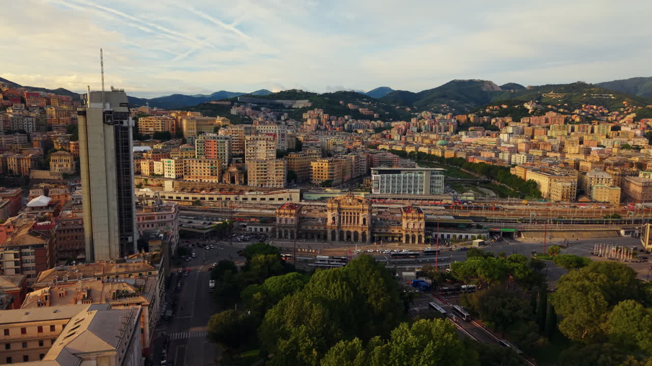 The drone pans above Genoa train station, showing a modern tower, surrounding buildings, and mountains in the background at sunset
