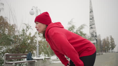 woman in red hoodie and beanie bends down resting, gazing into distant with snowy background featuring lamp pole, frosted trees, benches iron railing, and mast in a foggy atmosphere