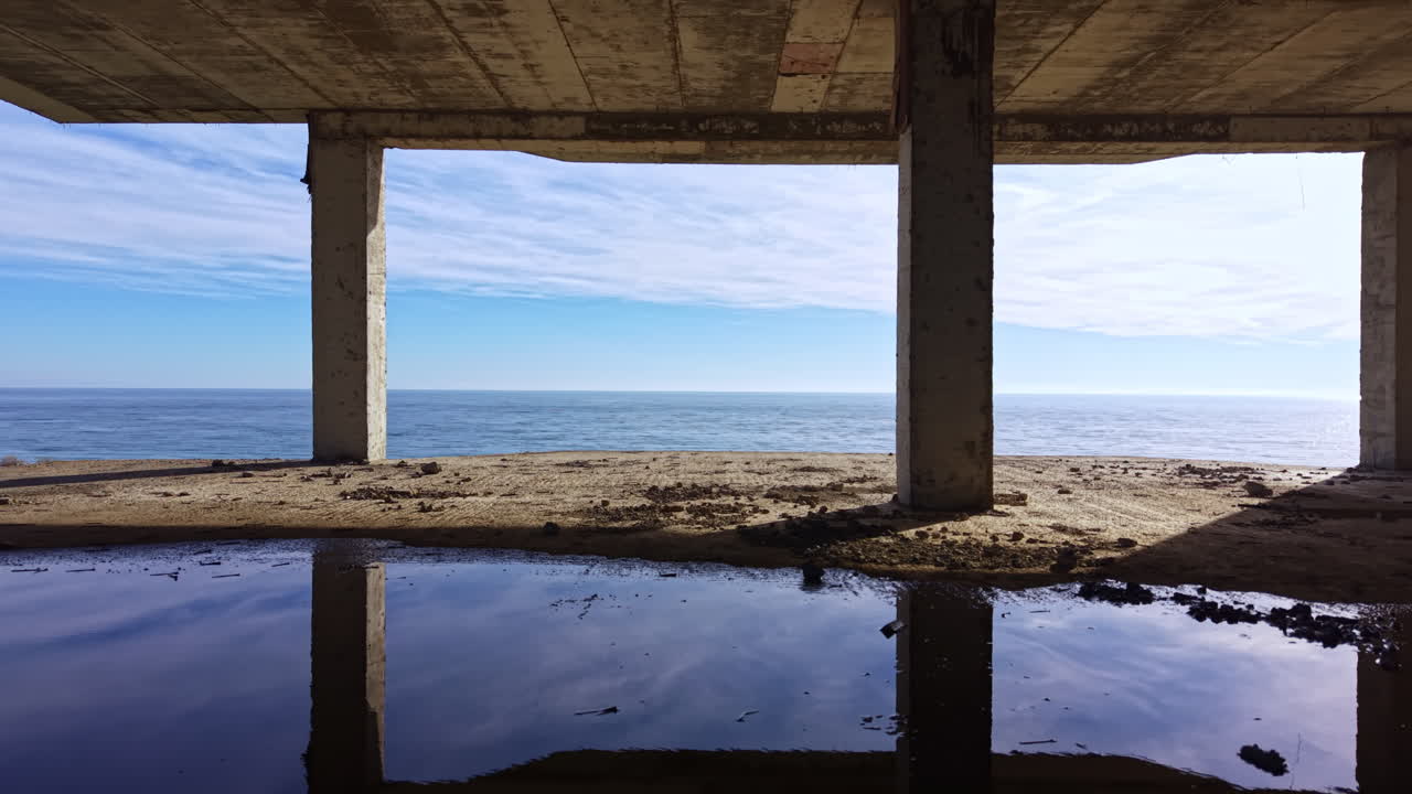 Serene coastal view from beneath an old structured bridge during a calm morning at the beach, offering a glimpse of clear skies and inviting waters