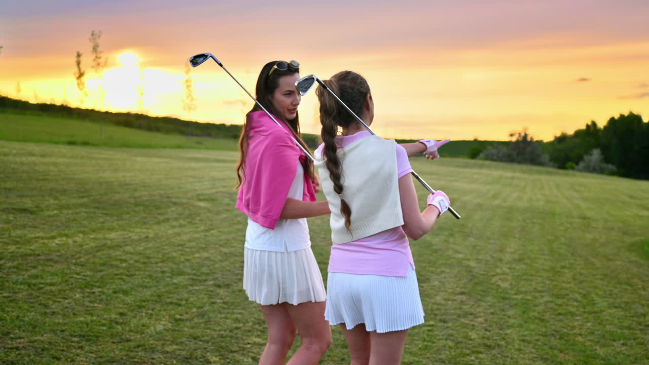 Due donne vestite con abiti bianchi e rosa, con mazze da golf sulle spalle, che camminano e parlano sul campo da golf