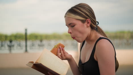 mujer con camiseta negra sentada al aire libre, disfrutando de un bocadillo mientras lee un libro, con una valla decorativa y un cielo brillante en el fondo, combinando casualmente la lectura y la comida
