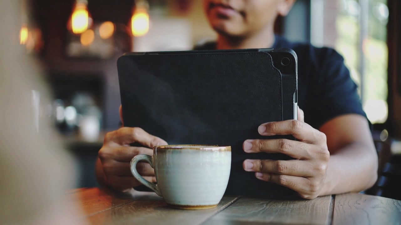 Uomo seduto con una camicia scura che sistema la custodia del tablet mentre legge a un tavolo del bar.