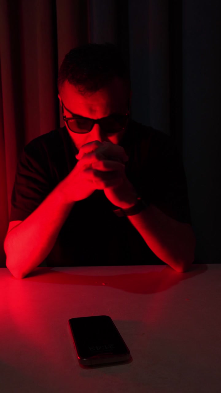 A dramatic medium close-up of a serious man wearing sunglasses talking on a smartphone in a dark room. The cinematic scene is intensely illuminated by vibrant red studio lighting.