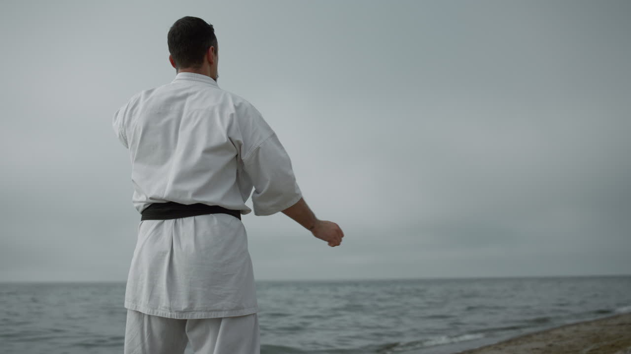 Atleta irriconoscibile si allena in posizioni di karate su una spiaggia sabbiosa in una giornata nuvolosa.