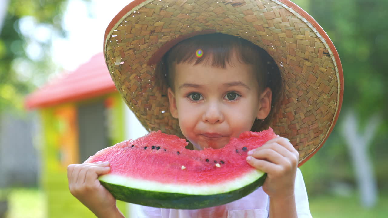 Lovely Caucasian kid wearing a big straw hat eating a watermelon. Sweet toddler enjoying ripe fruit outdoors. Close up. Blurred backdrop.