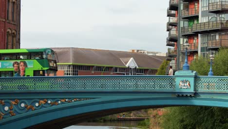 a green double-decker bus travels across a blue iron bridge in leeds, with modern buildings and overcast daylight. camera remains stationary, wide shot
