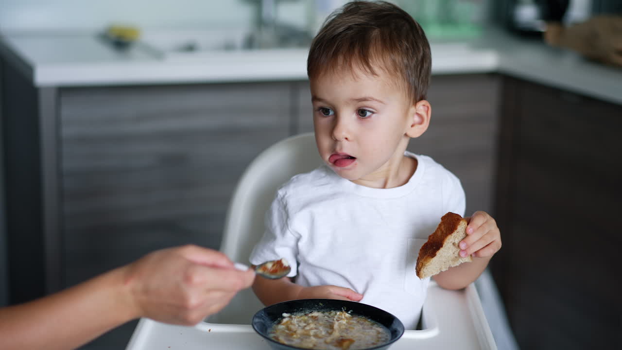 Mom's hand gives a spoon to her son. Toddler refuses to eat and waves his head. Blurred backdrop.