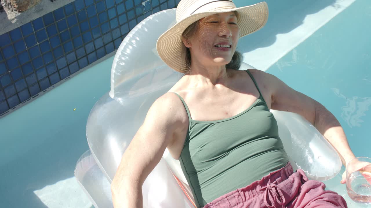 Settling into float, senior is reclining at pool edge wearing straw hat, holding glass, relaxing