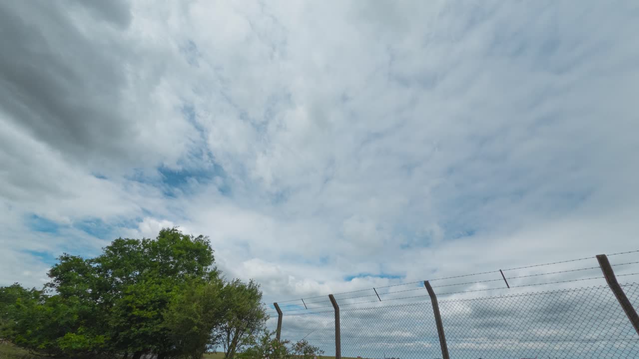 View of a cloudy sky with a wire fence, green landscape, and a distant building