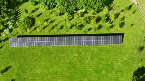 aerial view directly above a row of solar panels on a green field, surrounded by trees casting long shadows in the sunlight