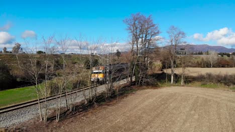 Aerial:-regional-train-in-Catalonia-emerging-from-trees-in-the-countryside-and-blue-sky