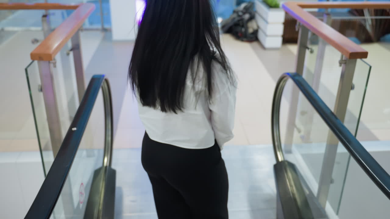 Confident woman in glasses and white shirt steps off escalator and glances back while smiling in bright modern mall environment with soft light and clear handrail reflections
