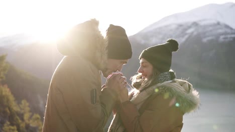 young couple in winter coats standingto each other, holding hands towards gorgeous snowy mountains and lake, a man warming his girlfiends hands and kissing. travel concept. sun strongly shining on the background