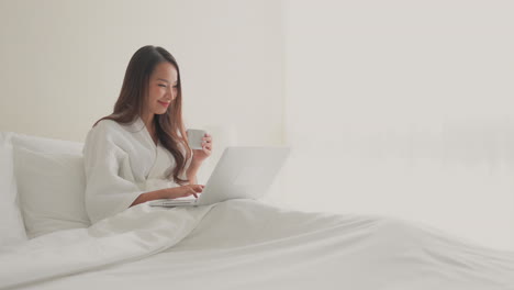 Toma-Amplia-De-Una-Joven-Asiática-Adulta-Con-Albornoz-Blanco-Sentada-En-Una-Cama-Doble-De-Hotel-Con-Una-Taza-Trabajando-En-Su-Laptop-Sonriendo-Alegremente-En-Una-Habitación-Blanca