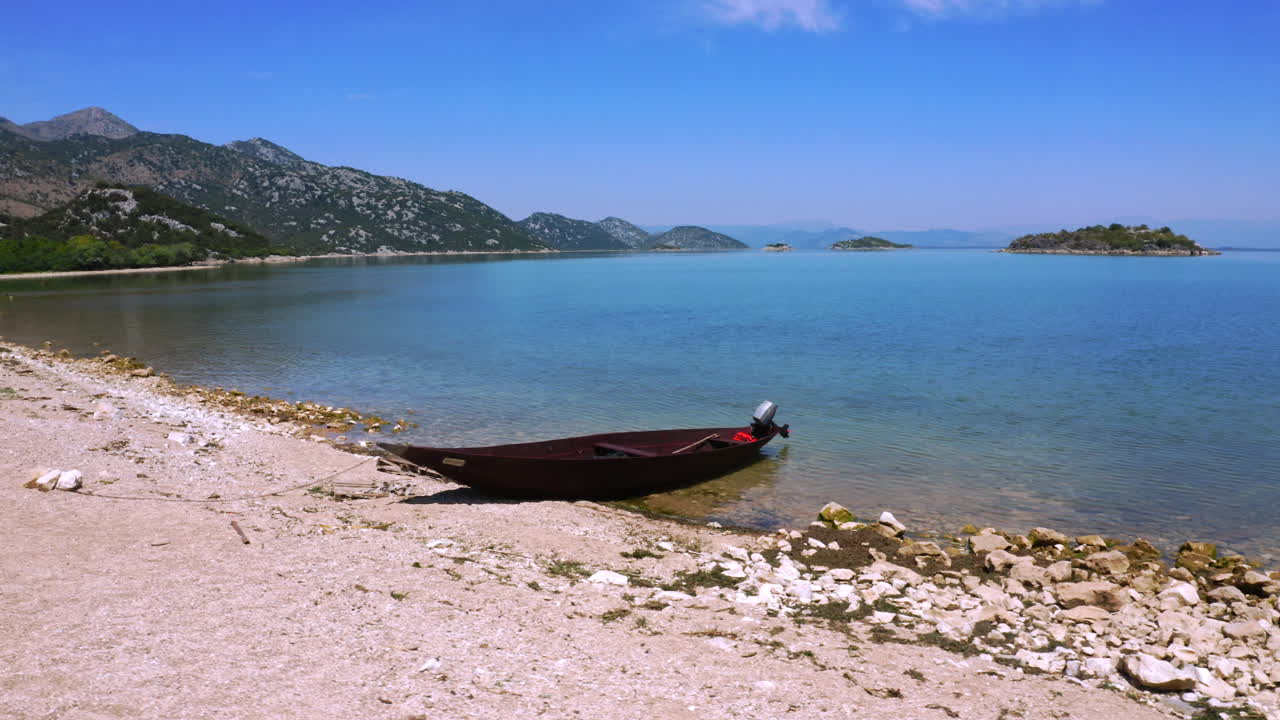 A small dark wooden motorboat anchored on the rocky beach of lake Skadar in Montenegro, flying birds above