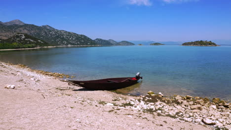 una pequeña lancha de madera oscura anclada en la playa rocosa del lago skadar en montenegro, pájaros volando arriba