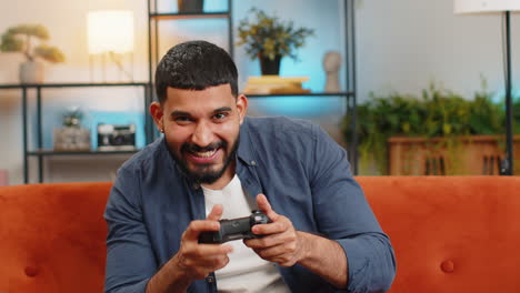 hombre indio usando controlador de joystick jugando a la consola de video ganando juego divertido en línea se sienta en el sofá de casa