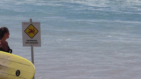 surfer walks by a warning sign towards the sea