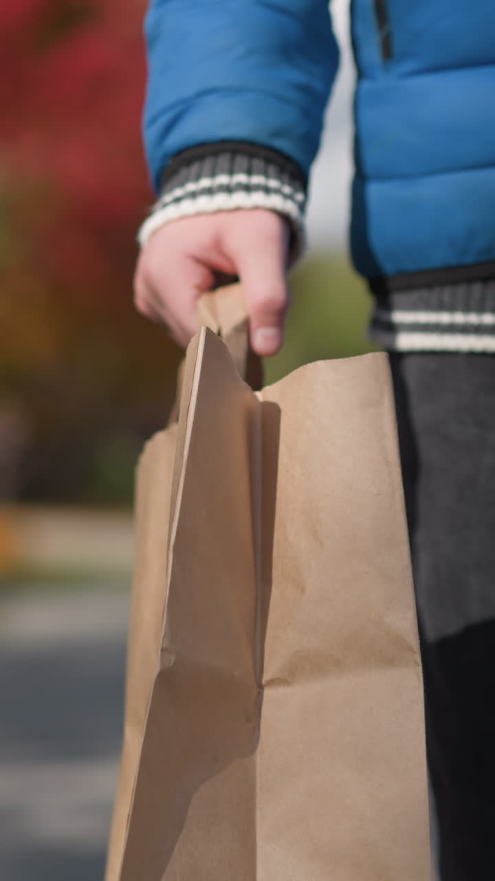 vista parcial de alguien caminando en una zona residencial llevando una bolsa de papel, enmarcada por árboles de otoño, casas y un coche estacionado en el fondo de enfoque suave