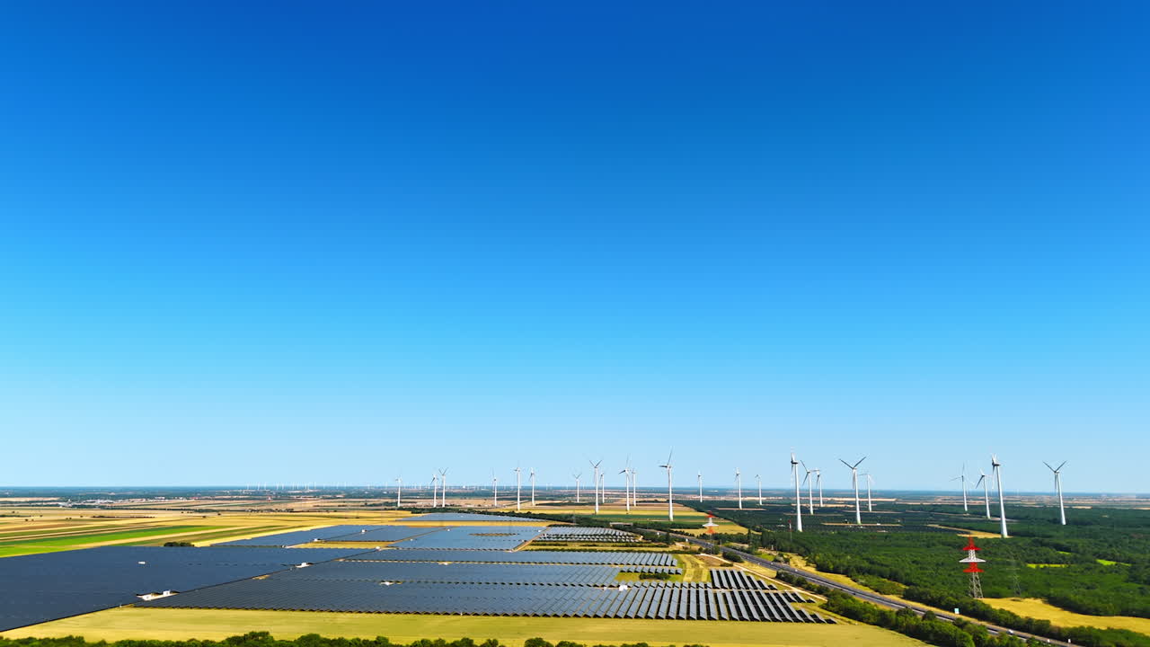 Approaching fields covered with multiple solar panels. Wind farms nearby produce green energy. Aerial view