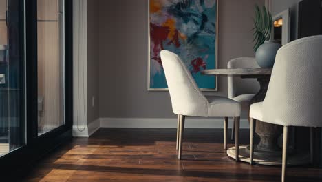 panning-shot-of-a-dining-table-setup-with-white-chairs,-a-circular-table,-colorful-artwork,-right-next-to-a-window-letting-in-natural-sunlight