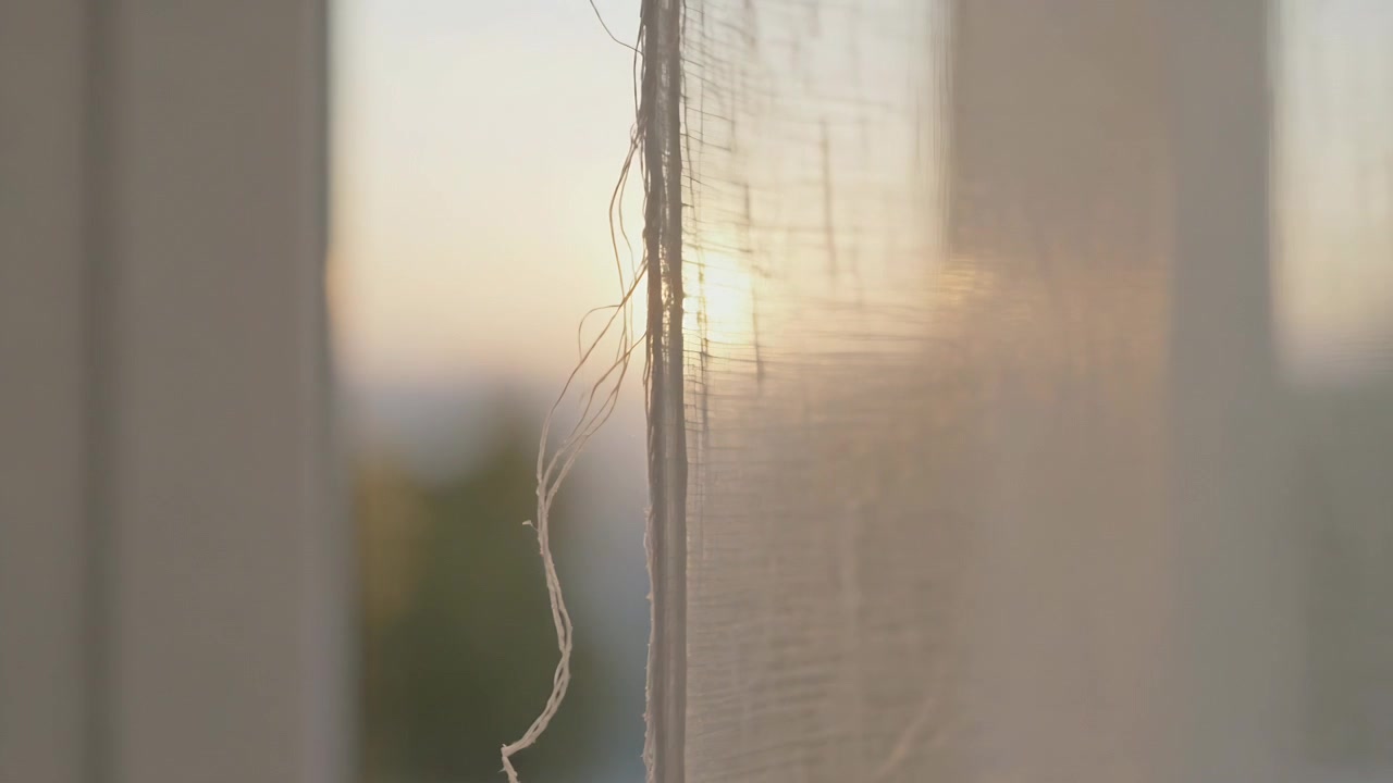 Il sole che si sposta, il bordo di una tenda velata con un filo allentato che brilla e si muove alla finestra, rivelando la trama.
