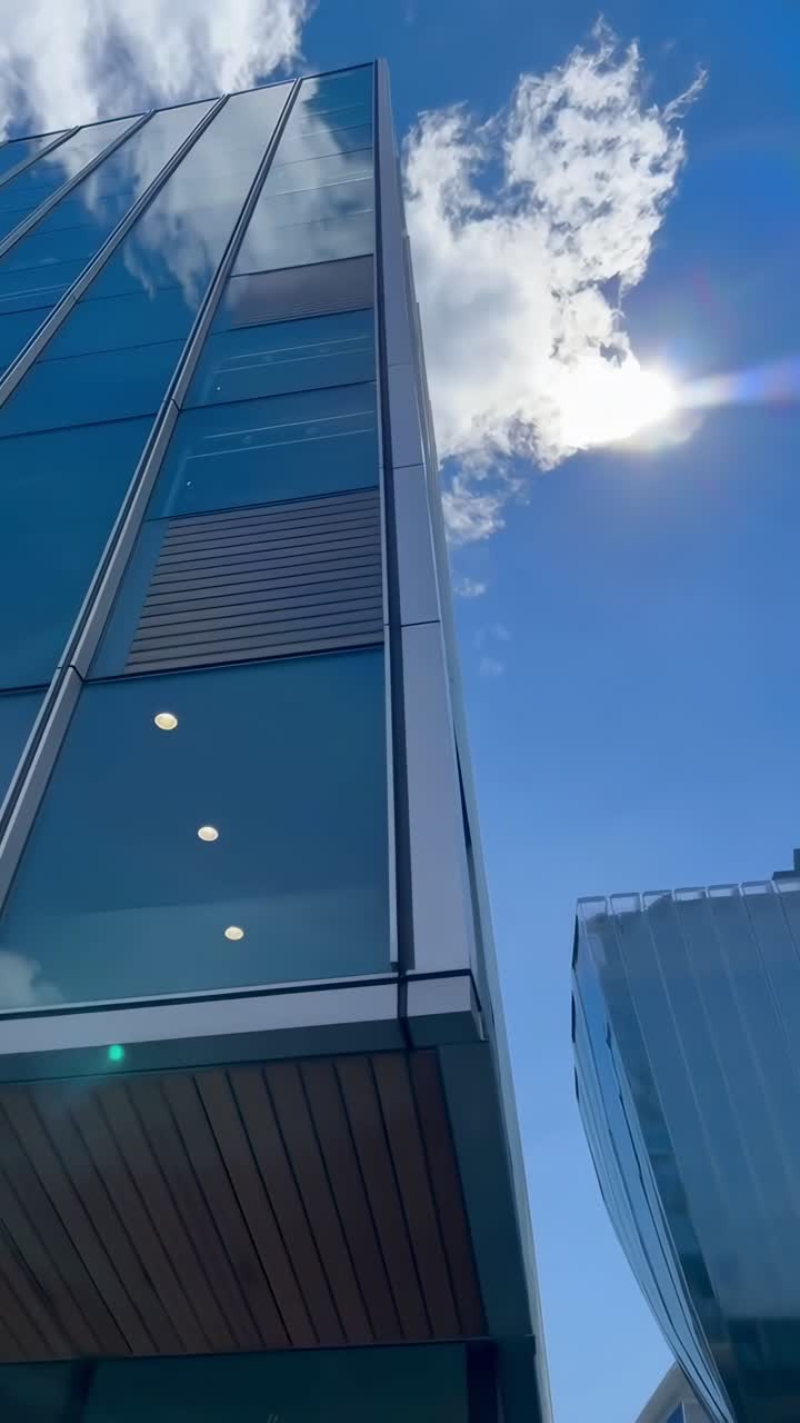 Video en contrapicado de un edificio moderno de cristal contra un cielo azul con nubes, enfatizando la altura.